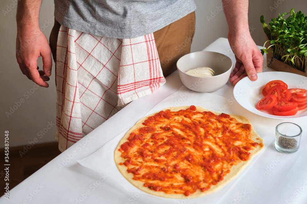 A man is preparing pizza. Cooking homemade italian pizza. Preparation raw ingredients for baking. Sauce, tomatoes and cheese. Fresh natural healthy food. Сulinary сhef kneading dough on kitchen table