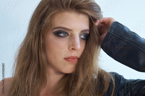 Natural beautiful young woman with light brown long hair and light gray eyes wearing a black leather jacket, resting her head on her arm while glancing thoughtfully into the distance.