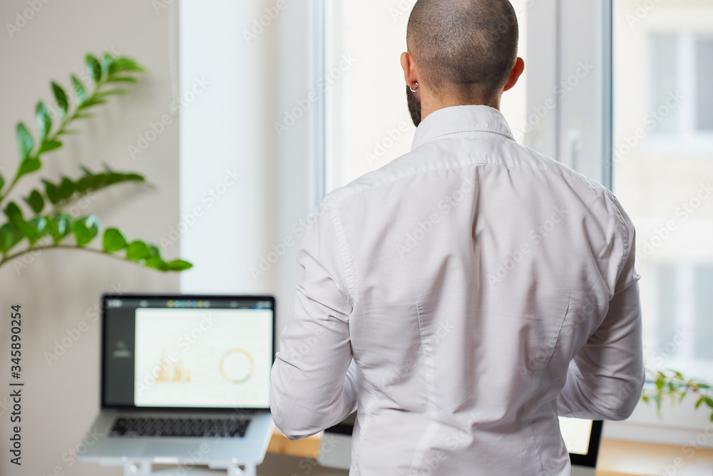A man with a beard in a white shirt standing with his back in the office with computers and green plants in the background.