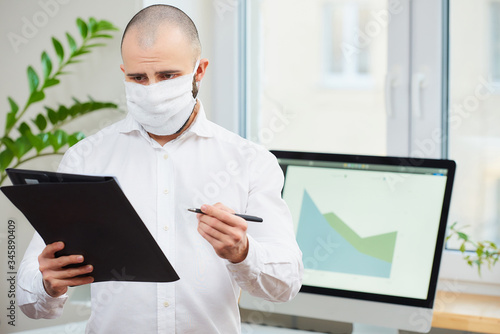  A man in a medical face mask against the coronavirus (COVID-19). An office worker holding a pen and a folder at his workspace with computers and green plants in the background. Coronavirus quarantine