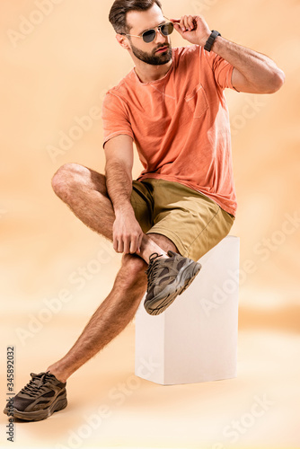 handsome young man in shorts, summer t-shirt and sunglasses sitting on white cube on beige photo