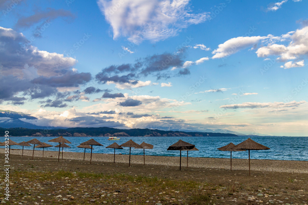 Straw beach umbrellas on the Empty Black Sea coast in Abkhazia, Pitsunda at sunset. Pebble beach, mountains, sea, sky with clouds Straw beach umbrellas on the Empty Black Sea coast in Abkhazia, Pitsunda at sunset. Pebble beach, mountains, sea, sky with clouds