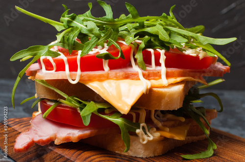 Very appetizing tasty big sandwich with ham or balyk, arugula, cheese, tomatoes and mayonnaise on a cutting board in a rustic style on a gray background view from side close-up, copy space
