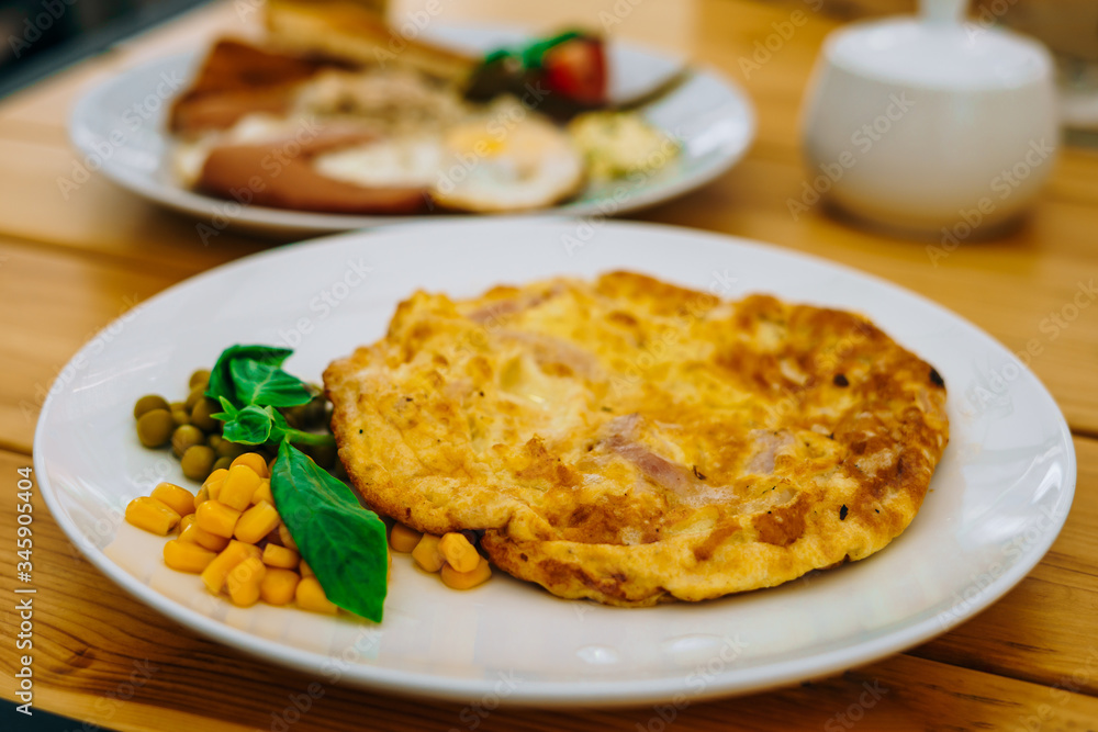 Omelet with ham, peas and corn in a cafe. Delicious omelet breakfast with cheese and peas. Omelet with vegetables on a white plate in a cozy cafe.