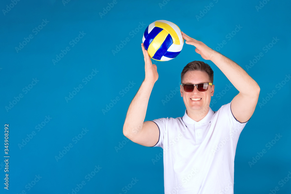 Naklejka premium A young volleyball fan, a man in a white shirt, stands with a ball in his hands on a blue background. The concept of recreation and travel.