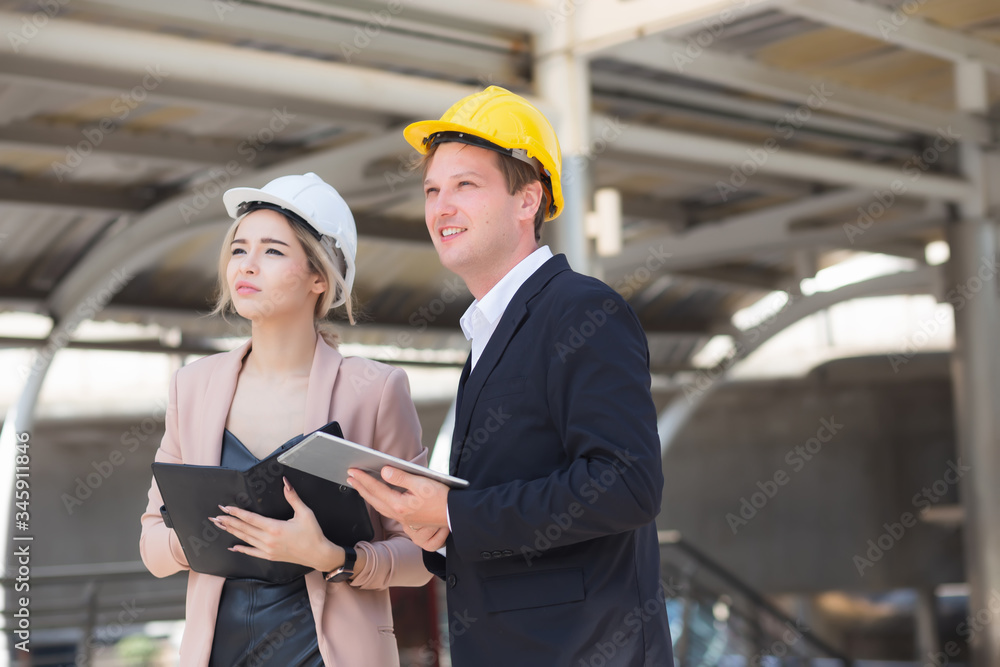 Male and female engineers wear formal suits and hard hats in mobile phones, documents, and tablets. They are looking at and checking the progress of the Sky Train project behind the station. Male and female engineers wear formal suits and hard hats in mobile phones, documents, and tablets. They are looking at and checking the progress of the Sky Train project behind the station.