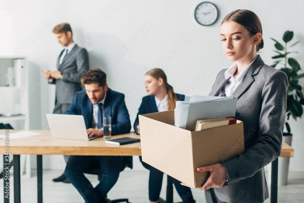 Dismissed woman holding cardboard box near colleagues working in office Dismissed woman holding cardboard box near colleagues working in office