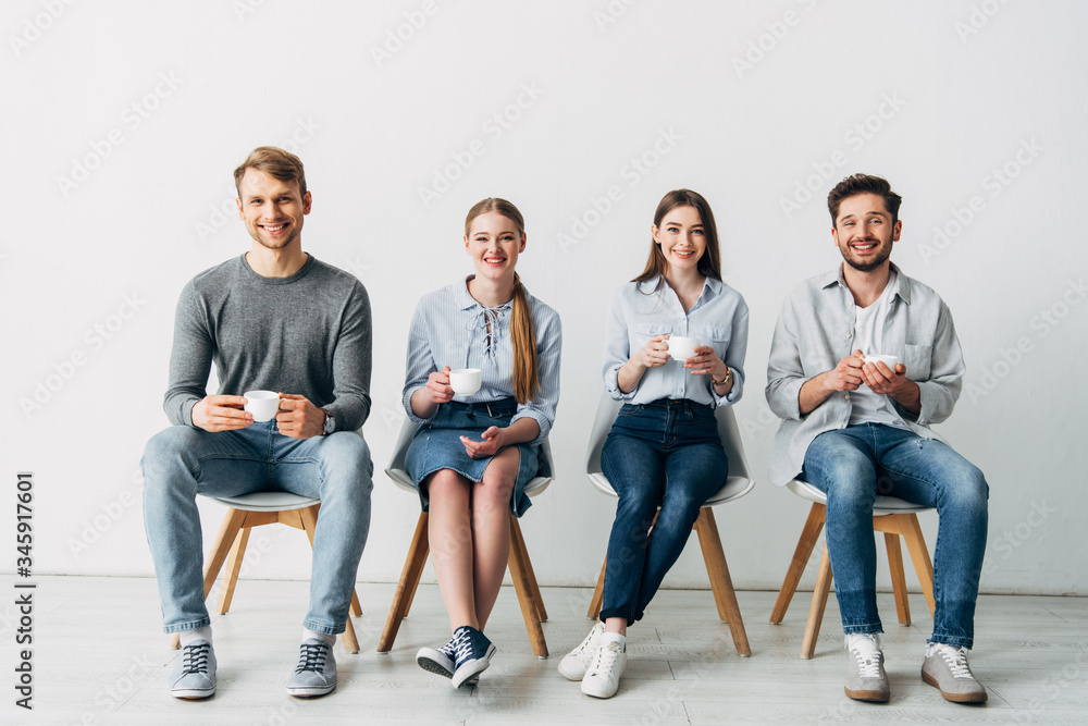 Smiling coworkers with coffee cups smiling at camera on chairs in office