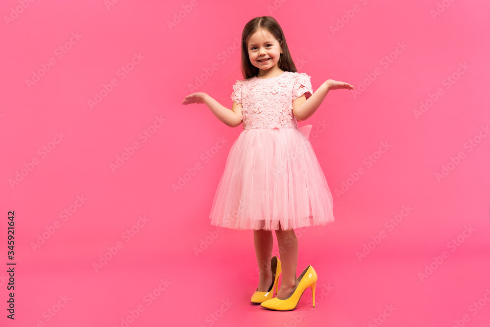 Amazing excited happy girl in dress in mama`s shoes expressing to camera isolated on pink background. Brightful emotions. Amazing excited happy girl in dress in mama`s shoes expressing to camera isolated on pink background. Brightful emotions.