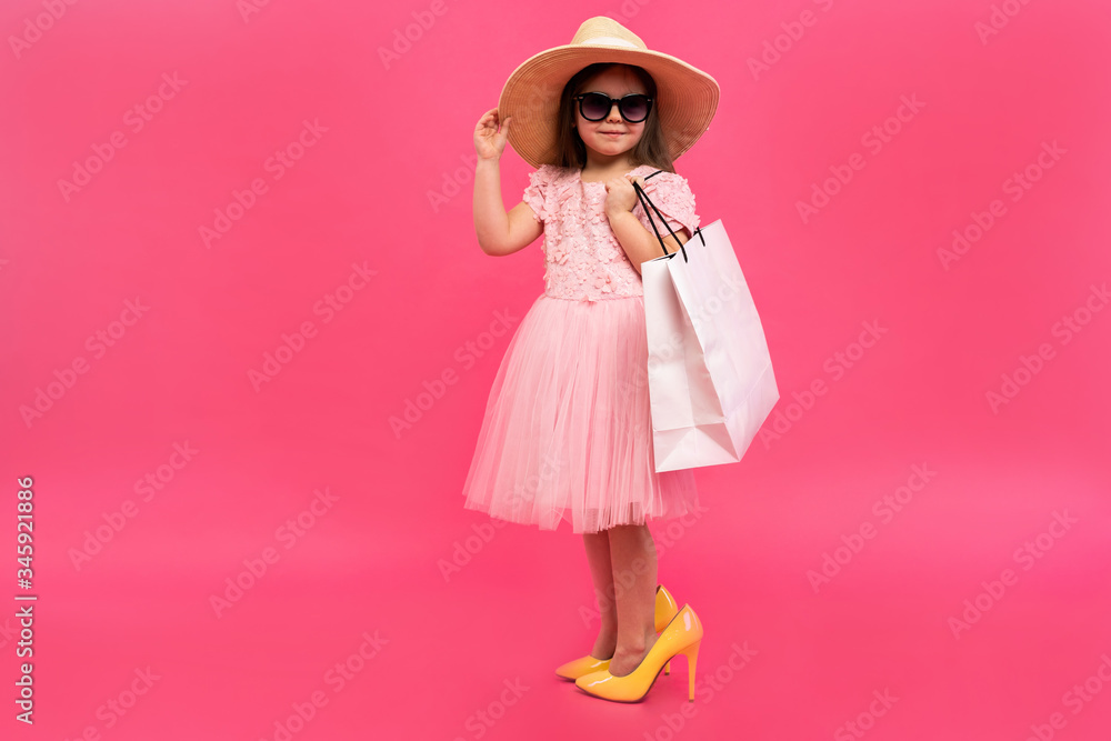 Happy lovely moments of shopping time with cute little girl in dress standing in mother`s big shoes with white packages in hands isolated on pink background Happy lovely moments of shopping time with cute little girl in dress standing in mother`s big shoes with white packages in hands isolated on pink background