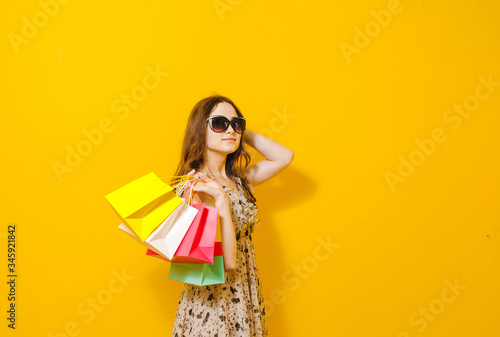 Portrait of a beautiful brunette girl smiling with sunglasses, holding shopping bags over yellow background