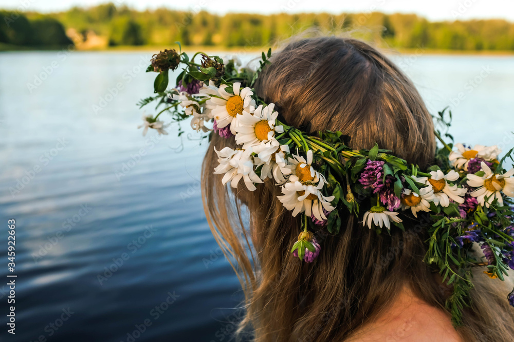 Naklejka premium Blonde girl in a white dress with a flower wreath on her head is sitting on the pier. Scenic forest lake. Ligo.