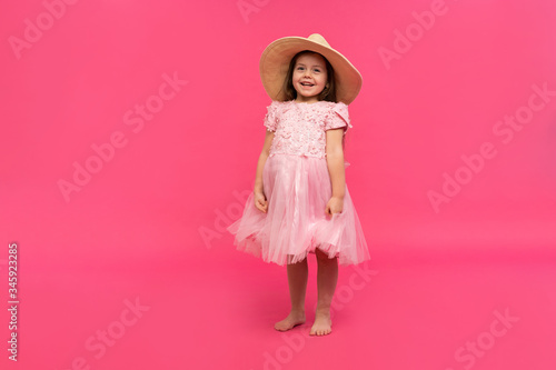 Portrait of cute little girl in straw hat and pink dress in the studio on pink background. Copy space for text.