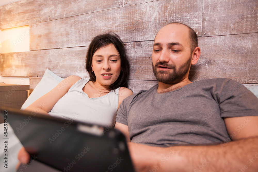 Happy boyfriend and girlfriend wearing pajamas lying in bed browsing on tablet computer. Happy boyfriend and girlfriend wearing pajamas lying in bed browsing on tablet computer.