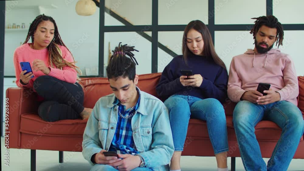 Group of young multiracial diverse friends using smartphone with mutual disinterest towards each other in loft appartment. Phone addicted young multiethnic people phubbing each other indoors. Group of young multiracial diverse friends using smartphone with mutual disinterest towards each other in loft appartment. Phone addicted young multiethnic people phubbing each other indoors.