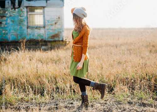 Fotografie Young woman wearing a dress, knitted cardigan and boots, posing in a field, backlit