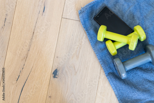 Fitness at home, equipment and tools for gym at home. Green dumbbell with towel, mobile phone on wooden parquet floor. Background with copyspace for sport activity at home with accessories.