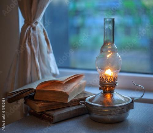 Vintage kerosene lamp with fire on a blurred background of the window and curtains with a book and glasses.