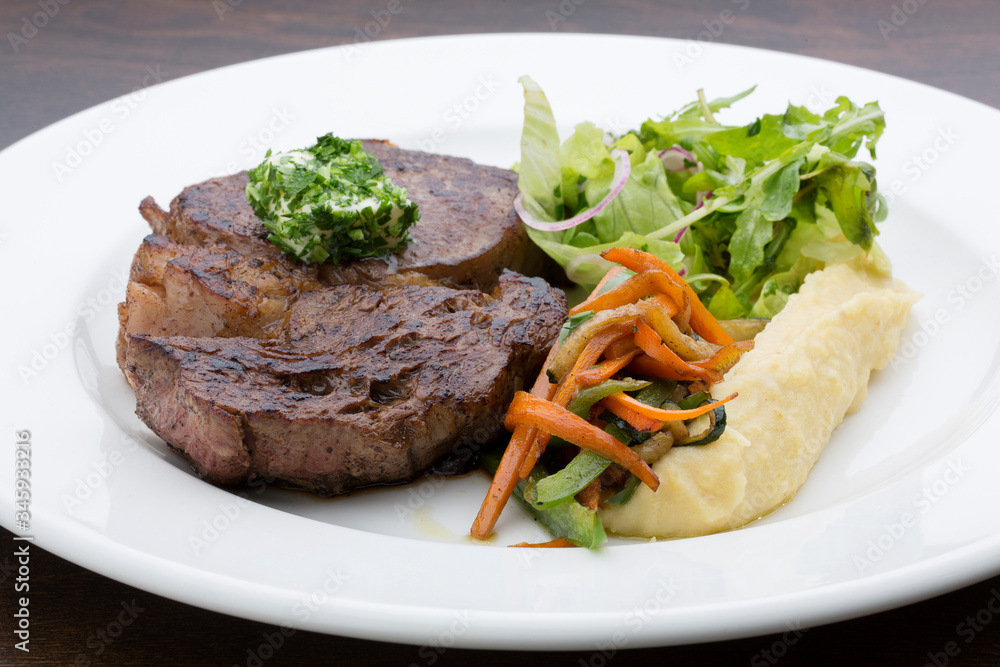 A roasted steak with mashed potatoes, fried vegetables and lettuce in a white ceramic plate on a wooden table