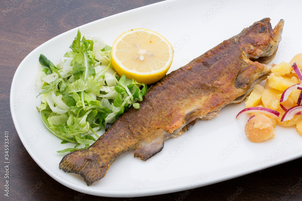 A roasted trout with potatoes, onions and lettuce in a white ceramic plate on a wooden table A roasted trout with potatoes, onions and lettuce in a white ceramic plate on a wooden table