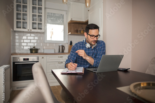 Businessman working remotely writing notes in his notebook while using laptop.