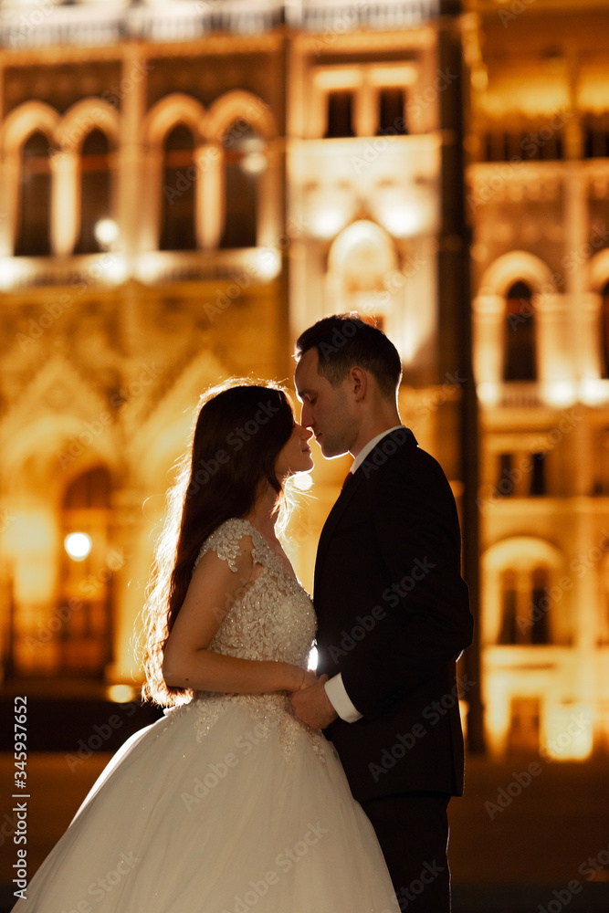 Fototapeta premium Bride and groom hugging in the old town street. Wedding couple walks in Budapest near Parliament House in night. Happy romantic young couple celebrating their marriage. Wedding and love concept.