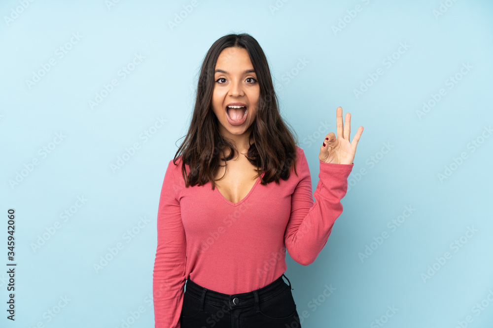 Young mixed race woman isolated on blue background surprised and showing ok sign Young mixed race woman isolated on blue background surprised and showing ok sign