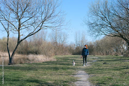 Woman wearing a protective mask is walking alone with a dog on country park road during coronavirus covid- 19