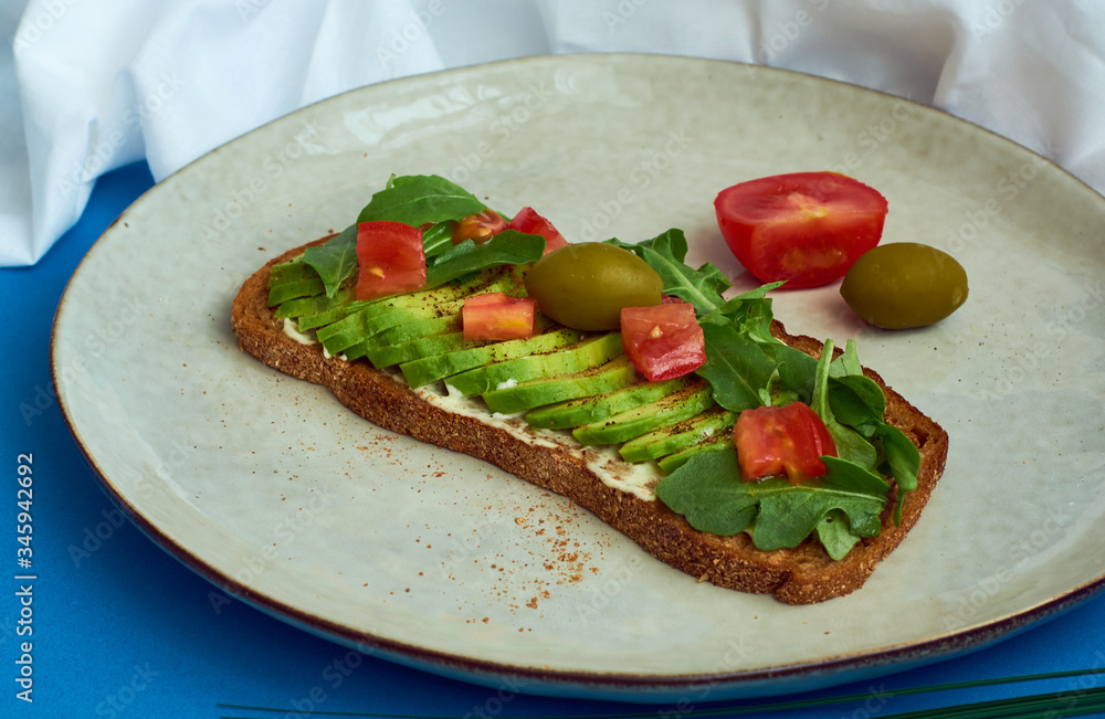 Avocado toast of rye bread with sauce, tomatoes, arugula on large grey plate on blue background. Vegetarian useful breakfast.