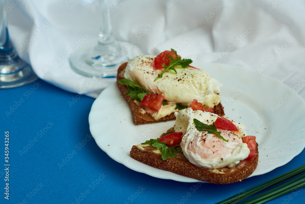 Poached eggs on sandwich of rye bread with sauce, tomatoes and arugula in white plate on blue background. Poached eggs on sandwich of rye bread with sauce, tomatoes and arugula in white plate on blue background.