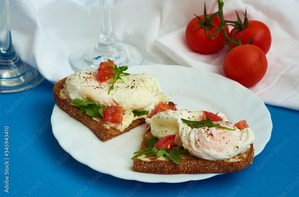 Poached eggs on sandwich of rye bread with sauce, tomatoes and arugula in white plate on blue background. Poached eggs on sandwich of rye bread with sauce, tomatoes and arugula in white plate on blue background.