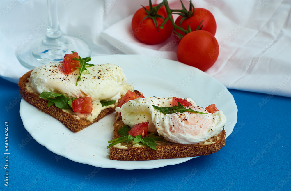Poached eggs on sandwich of rye bread with sauce, tomatoes and arugula in white plate on blue background. Poached eggs on sandwich of rye bread with sauce, tomatoes and arugula in white plate on blue background.