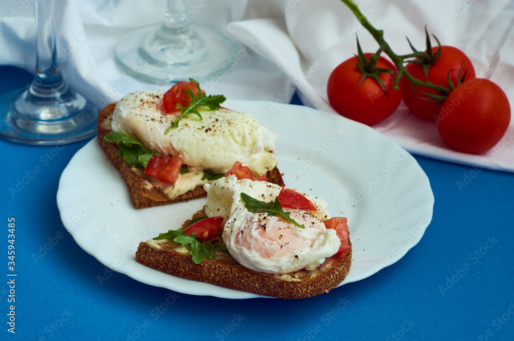 Poached eggs on sandwich of rye bread with sauce, tomatoes and arugula in white plate on blue background. Poached eggs on sandwich of rye bread with sauce, tomatoes and arugula in white plate on blue background.