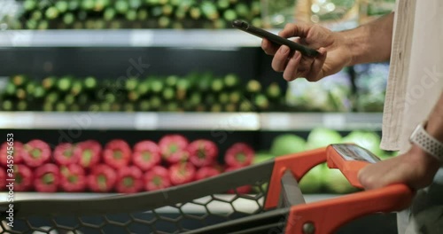 Crop view of man hand holding smartphone and srolling screen in supermarket. Guy using smartphone while walking and pushing shopping cart near vegetables shelves. Blurred background