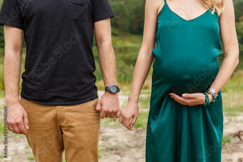 Pregnant woman in green dress and her husband in black t-shirt holding hands. Close up photo of pregnancy. Future mother touch belly with baby. Stylish interesting couple on the background of nature.