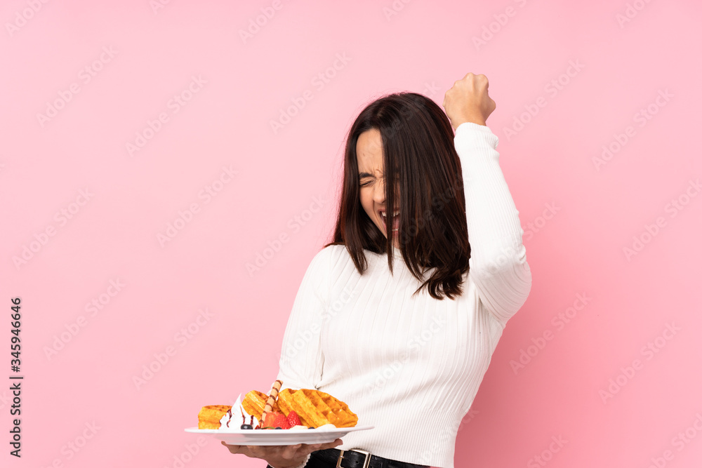 Young brunette woman holding waffles over isolated pink background celebrating a victory