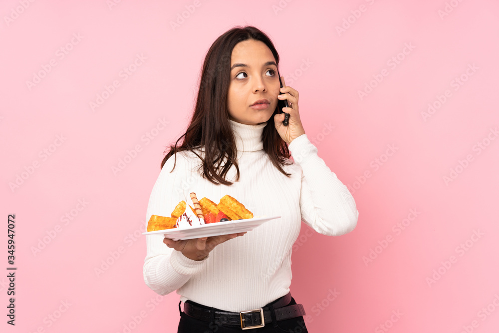 Young brunette woman holding waffles over isolated pink background holding coffee to take away and a mobile Young brunette woman holding waffles over isolated pink background holding coffee to take away and a mobile