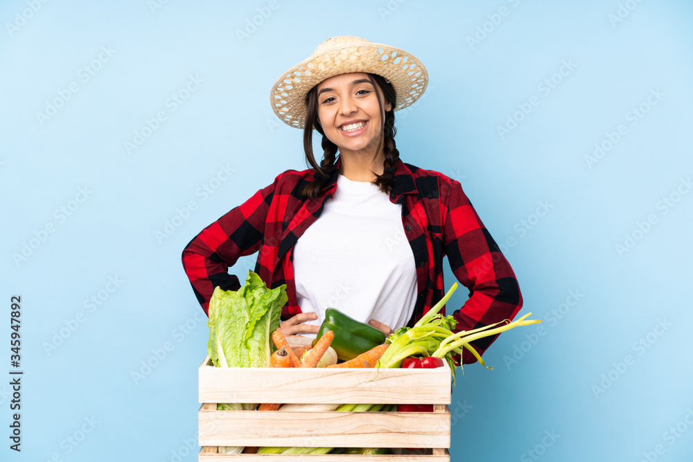 Young farmer Woman holding fresh vegetables in a wooden basket posing with arms at hip and smiling