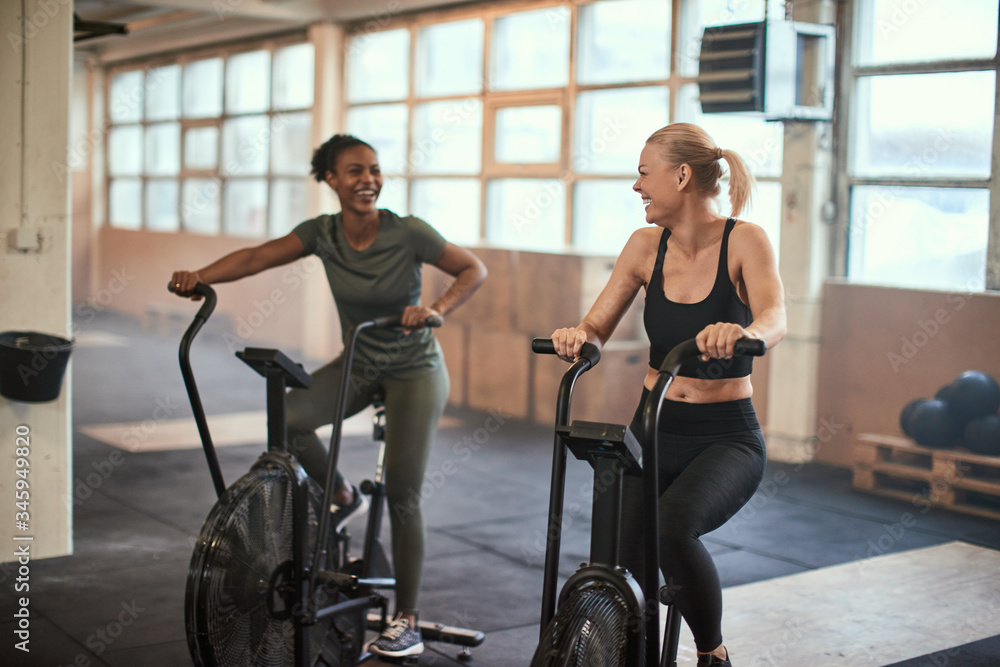 Fototapeta premium Two laughing young woman riding stationary bikes in a gym