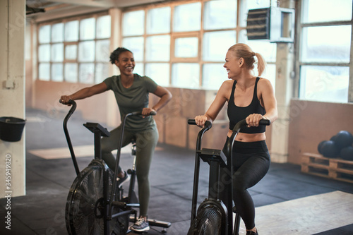 Two laughing young woman riding stationary bikes in a gym