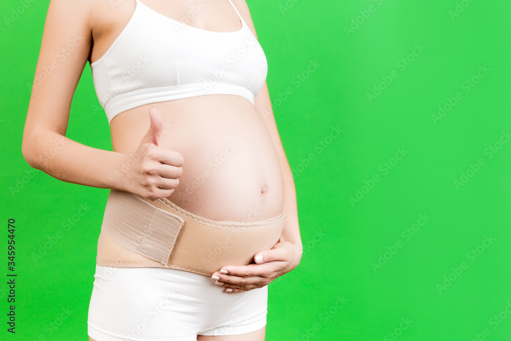 Close up of pregnant woman in underwear showing thumb up gesture and wearing maternity belt to reduce pain in the back at green background with copy space. Orthopedic abdominal support belt concept Close up of pregnant woman in underwear showing thumb up gesture and wearing maternity belt to reduce pain in the back at green background with copy space. Orthopedic abdominal support belt concept