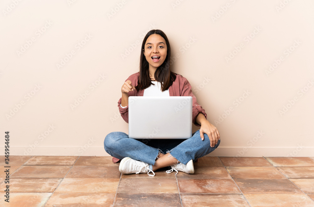 Young mixed race woman with a laptop sitting on the floor surprised and pointing front Young mixed race woman with a laptop sitting on the floor surprised and pointing front