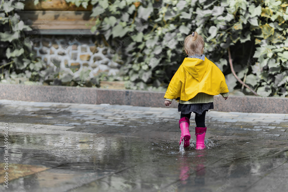 A nice little girl in a yellow raincoat and pink rubber boots jumps on puddles with splashes and rejoices. Park, nature, outdoors. Summer. Universal Children's Day.