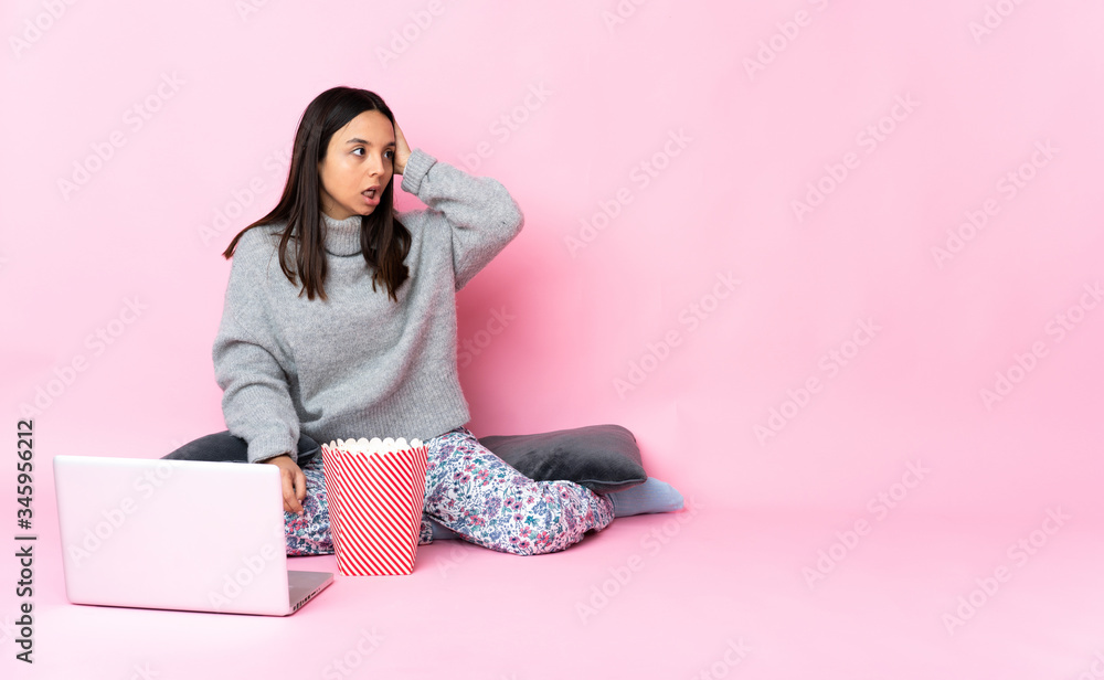 Young mixed race woman eating popcorn while watching a movie on the laptop doing surprise gesture while looking to the side Young mixed race woman eating popcorn while watching a movie on the laptop doing surprise gesture while looking to the side