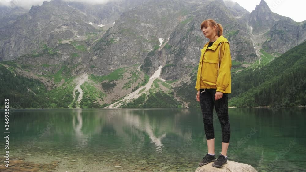 A tourist woman is standing on a stone at the shore of a beautiful mountain lake and looking at mountain peaks and then smiles into camera. Travel and adventure concept slow motion front view 4K video A tourist woman is standing on a stone at the shore of a beautiful mountain lake and looking at mountain peaks and then smiles into camera. Travel and adventure concept slow motion front view 4K video