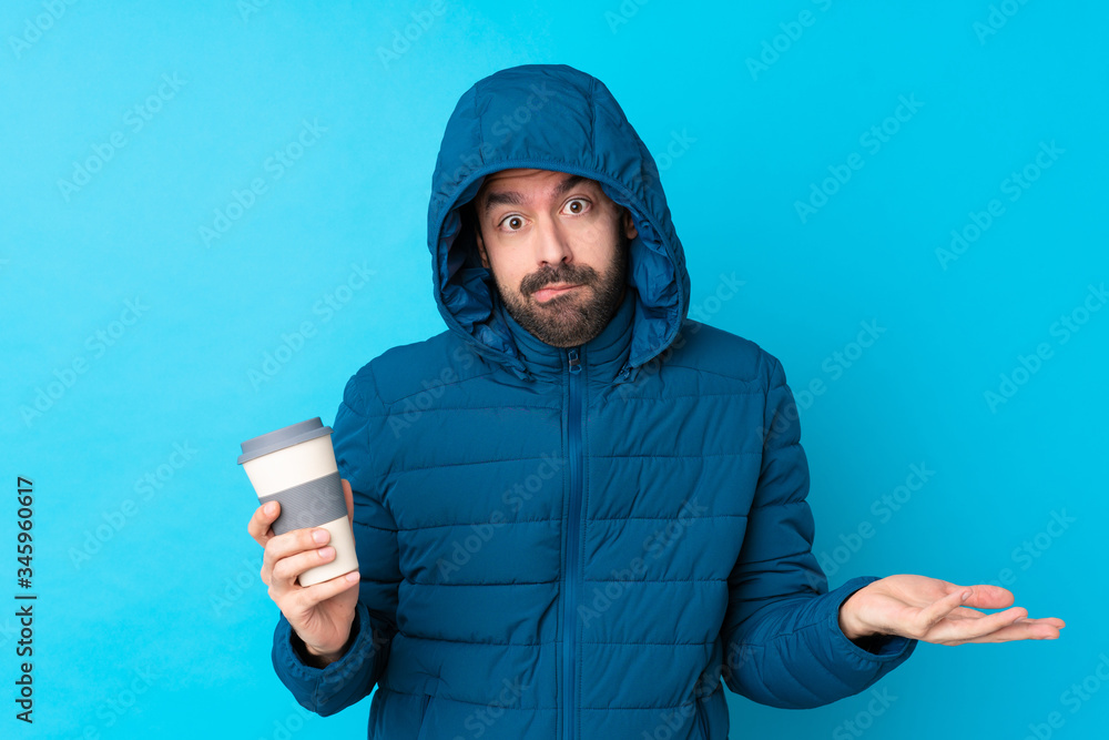Man wearing winter jacket and holding a takeaway coffee over isolated blue background having doubts while raising hands Man wearing winter jacket and holding a takeaway coffee over isolated blue background having doubts while raising hands