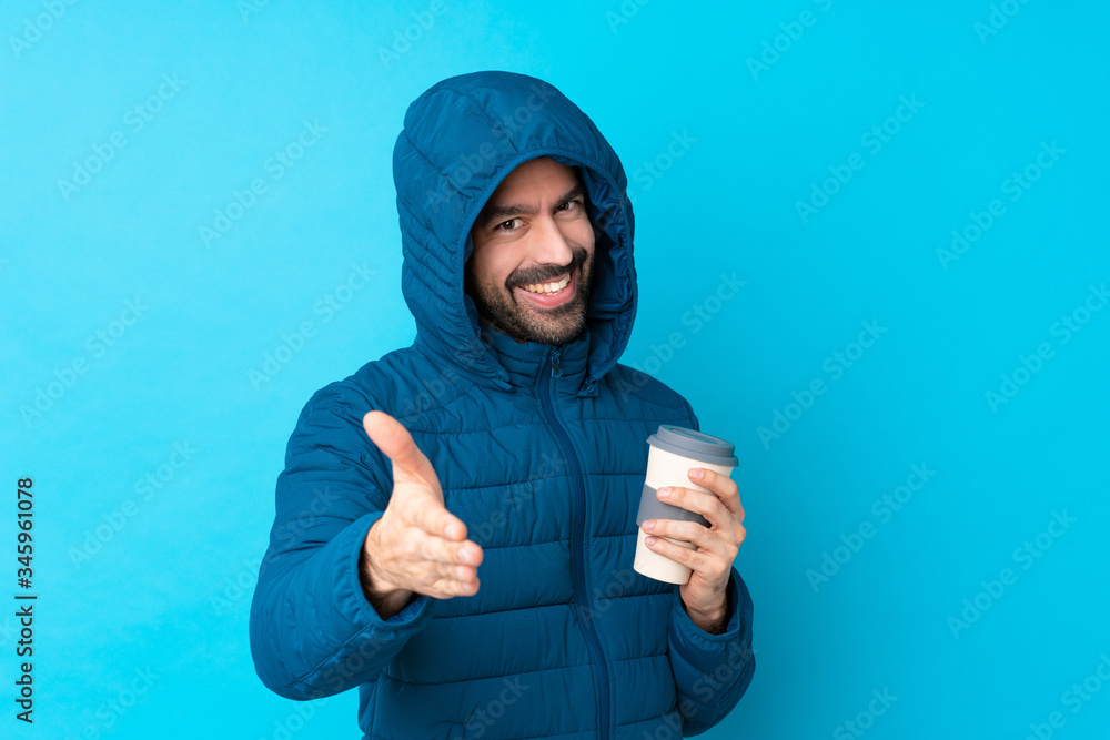 Man wearing winter jacket and holding a takeaway coffee over isolated blue background shaking hands for closing a good deal Man wearing winter jacket and holding a takeaway coffee over isolated blue background shaking hands for closing a good deal