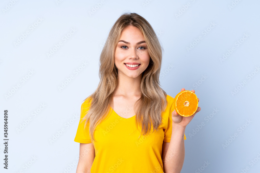 Teenager Russian girl holding an orange isolated on blue background smiling a lot Teenager Russian girl holding an orange isolated on blue background smiling a lot