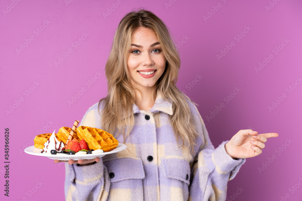 Teenager Russian girl holding waffles isolated on purple background pointing finger to the side Teenager Russian girl holding waffles isolated on purple background pointing finger to the side