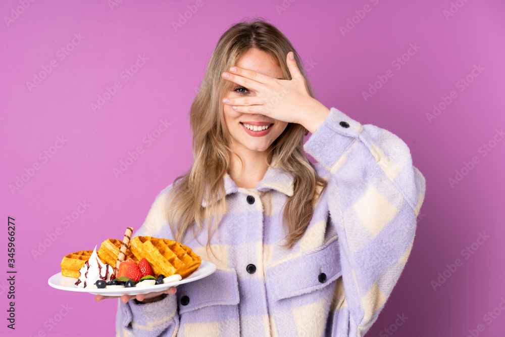 Teenager Russian girl holding waffles isolated on purple background covering eyes by hands and smiling